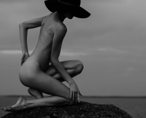 Wife, face hidden by hat, kneels nude on a large stone.