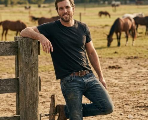 Handsome man leans against fence post on horse ranch ~ MarriageHeat